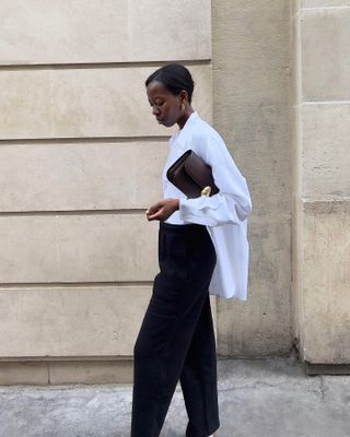 Fashion influencer Sylvie Mus poses on a Paris sidewalk wearing an open white button-down shirt, a white top, a burgundy clutch bag, and black pants.