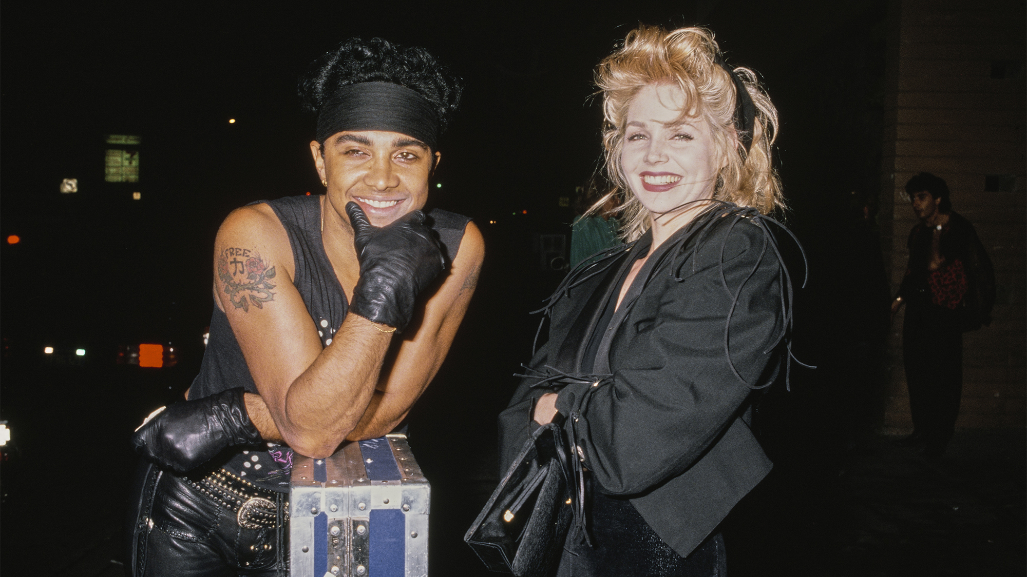 American singer and guitarist Micki Free, wearing black leather trousers with a black sleeveless top, his chin resting in his black-gloved hands, his elbows resting on a guitar case, as he stands beside American actress Teri Copley, who wears a black outfit, United States, circa 1985.