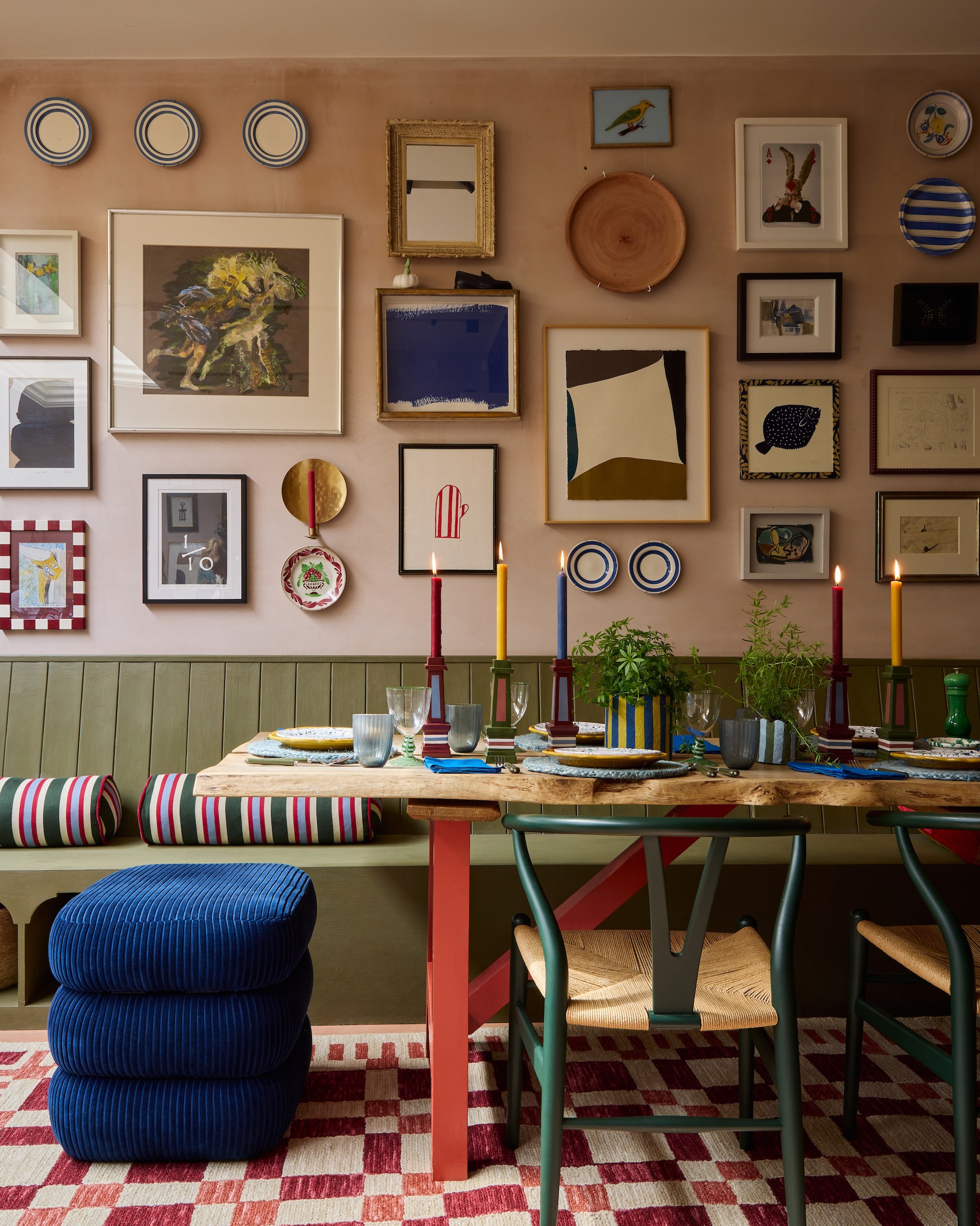 Dining space with a wood table and olive green, bench seat against a blush pink wall, covered in a collection of framed artwork.