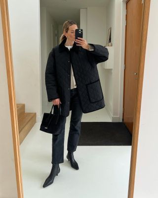 style influencer Anouk Yve poses for a mirror selfie wearing a quilted barn coat, cardigan, small black suede tote bag, black cropped ankle jeans, and black kitten heel boots