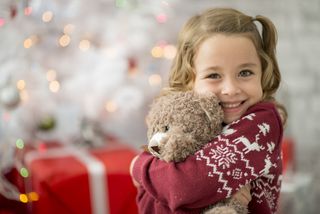 A young girl is indoors in a living room on Christmas day. She is wearing festive clothing. She is sitting in front of a Christmas tree and hugging her new teddy bear.