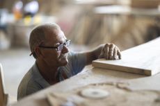 An older man, wearing glasses and with a pencil behind his ear, is measuring wood. He appears to be a working carpenter.