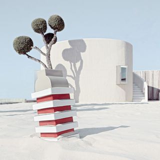 Daytime, outside image, white curved stone wall building and steps, small topiary tree in a slanted red and white striped pot, white sand, clear blue sky