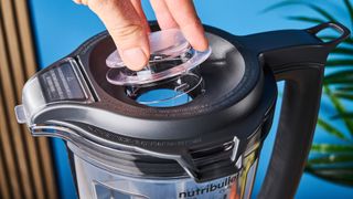 a black nutribullet with a big blending component is photographed in front of a blue backdrop, showing its plastic pitcher jug, silver blades, and tamper