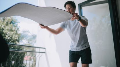 Man in shorts and T-shirt unfurling exercise mat in midair. He is outside on a balcony or porch