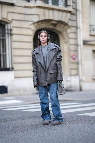 A woman wears a gray top, gray leather jacket, blue puddle jeans, and gray Ugg boots.