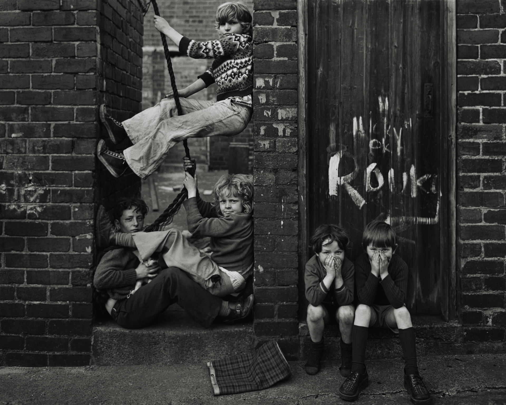 Black and white photograph of children playing on a rope swing wedged between two brick walls, with two more boys crouched nearby covering their faces, taken in Byker, Tyneside, in 1976.