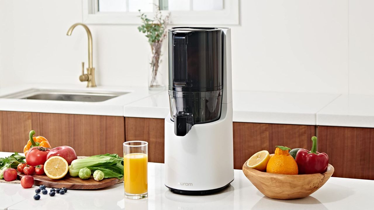 Hurom juicer in white on a white countertop with fruits to the left and right of it