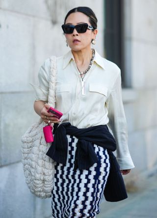 A woman in New York wearing a cream blouse, black sunglasses, Cos quilted bag and patterned skirt.