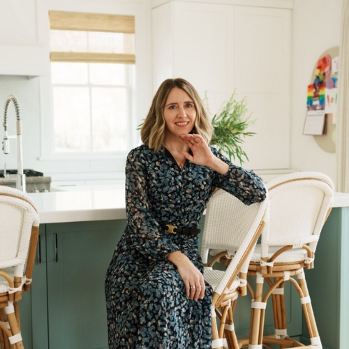 Image of a woman in a blue and black patterned dress sitting on a white rattan kitchen bar stool in a green and white kitchen.