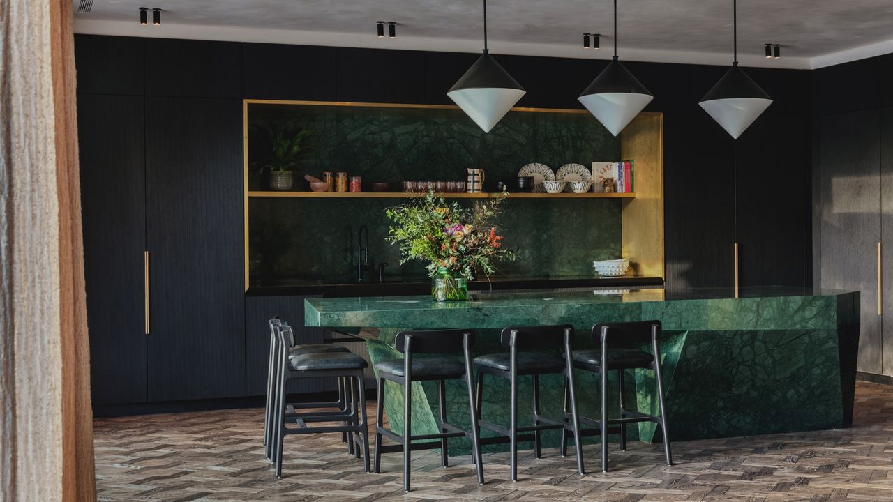 Image of a kitchen that has dark navy cabinetry. The built-in shelving has backsplash in a dark green marble that matches the material of the island. There are three black-and-white pendant lights hanging above the island and several minimal black stools positioned around one corner.