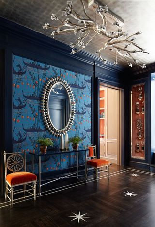 hallway with blue patterend wallpaper and orante mirror and ceiling light and decorative orange accent chairs