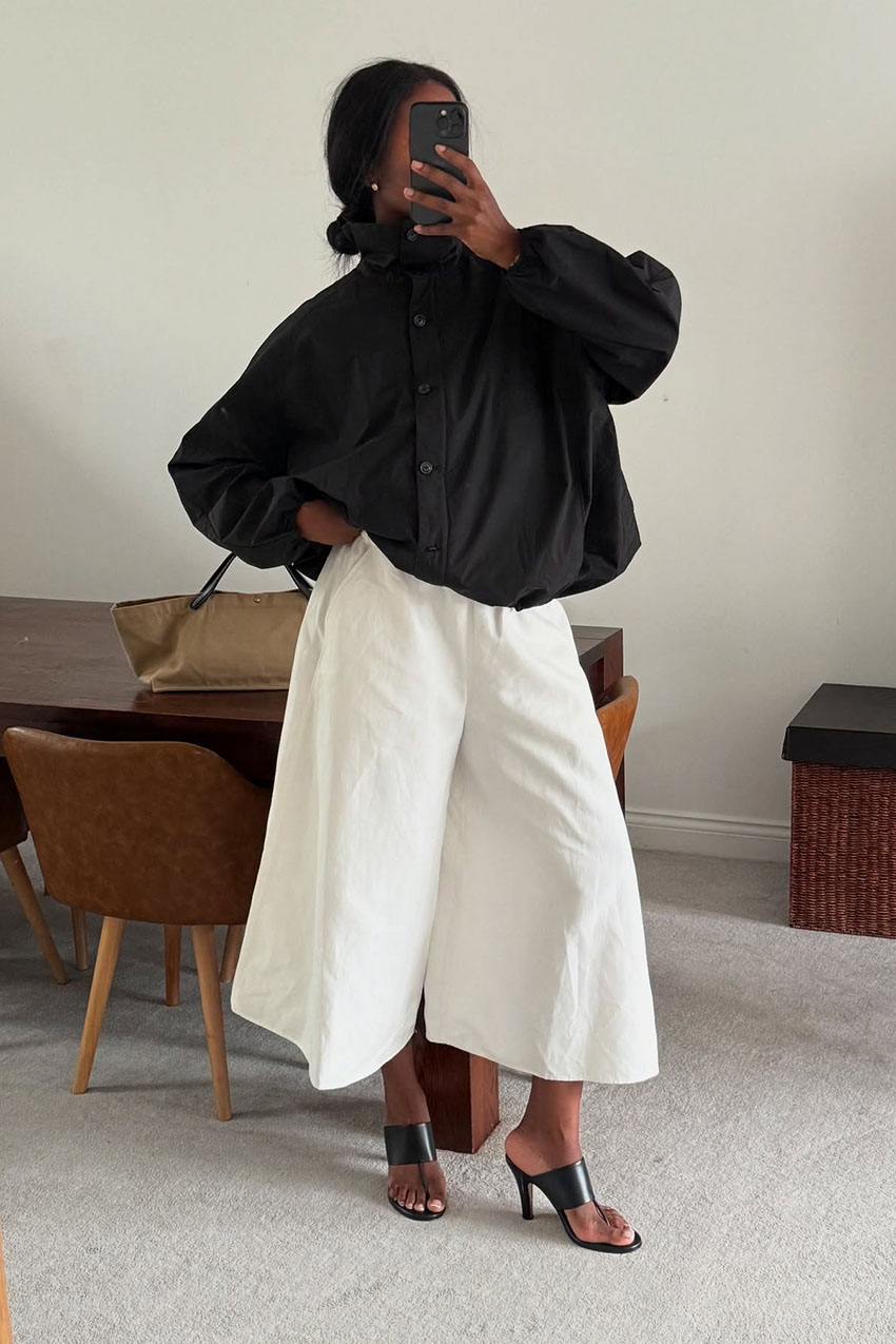 a woman wearing a black high collar jacket, white wide-leg pants, and black mule sandals