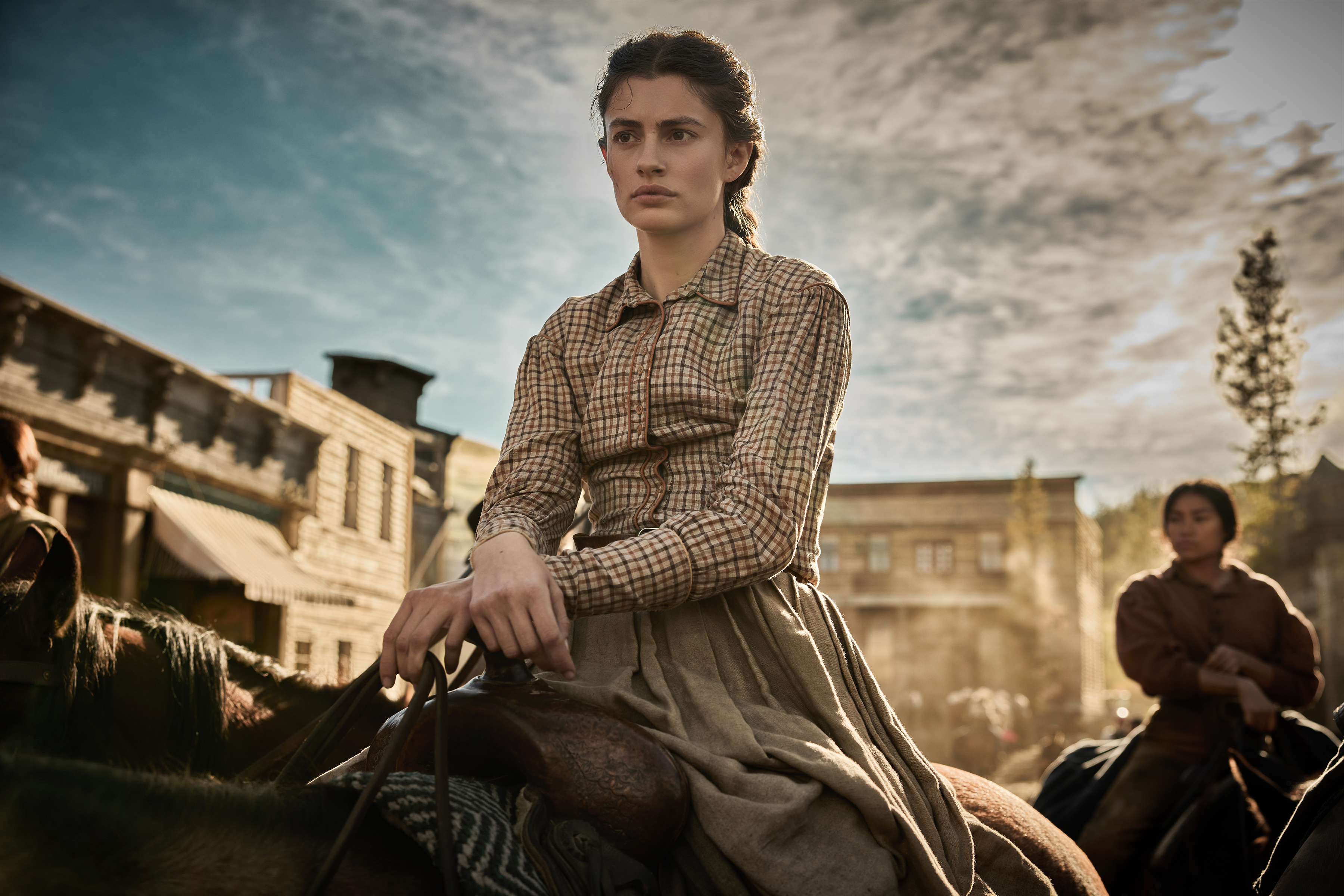 Diana Silvers as Dahlia Teller in The Abandons waering a plaid shirt and long skirt riding a horse through a ranch town