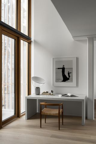 Modern residential interior featuring sleek white office desk and wooden chair to compliment the wooden framed windows, pale walls and light wooden floor.