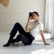 British influencer Jessica Skye poses for a mirror selfie wearing a basic white tee, western-inspired buckled belt, black raw-hem jeans, and black pointed-toe heeled boots