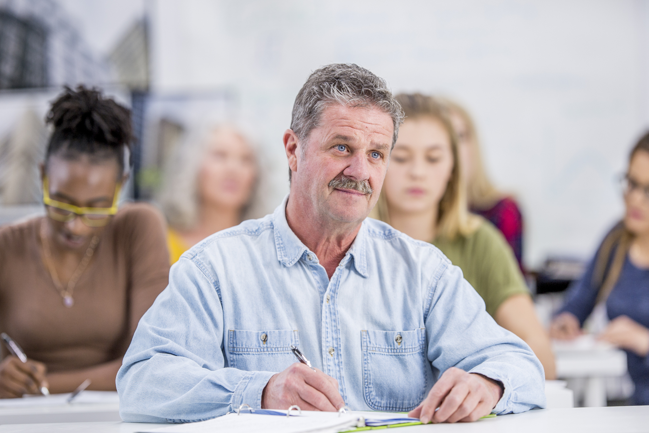 A group of students are indoors in a university. They are sitting during a lecture. A Caucasian man is in front, and he is listening to the professor.