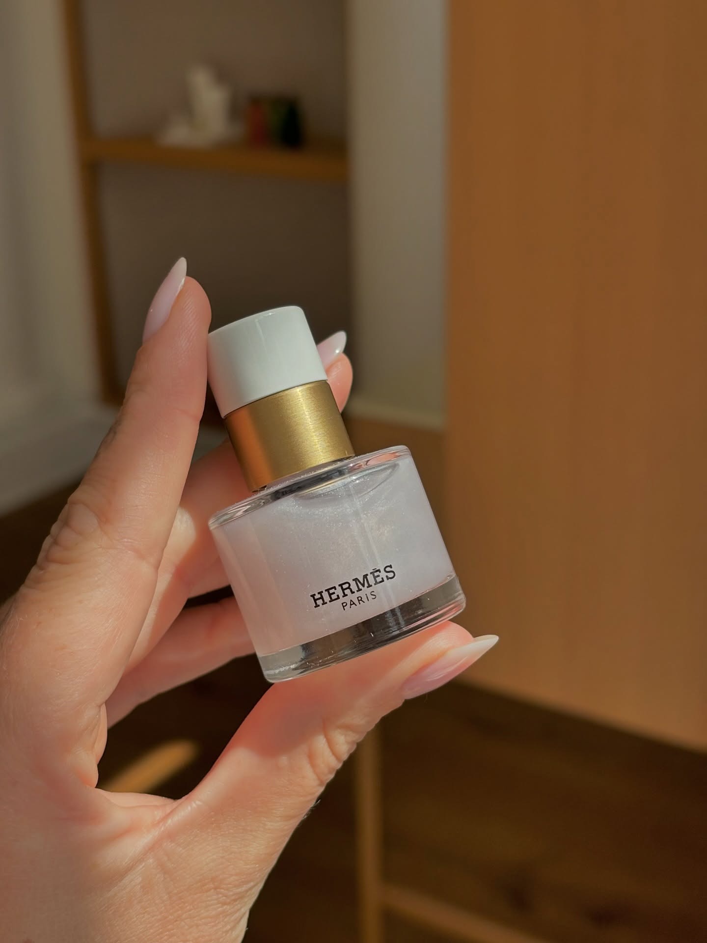 A hand holding a bottle of Hermes nail polish with a sheer shimmery manicure