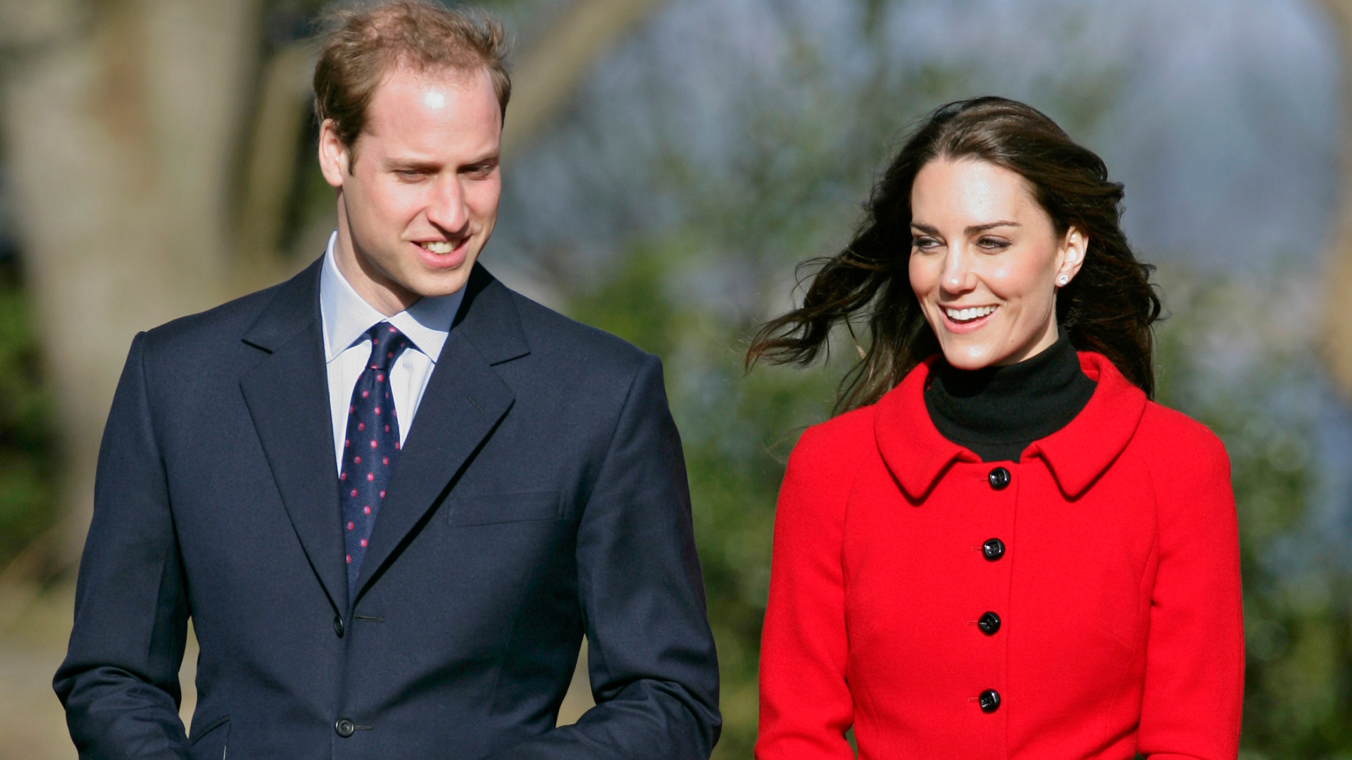 Prince William wears a suit and Kate Middleton wears a bright red coat with black buttons and a black polo neck underneath