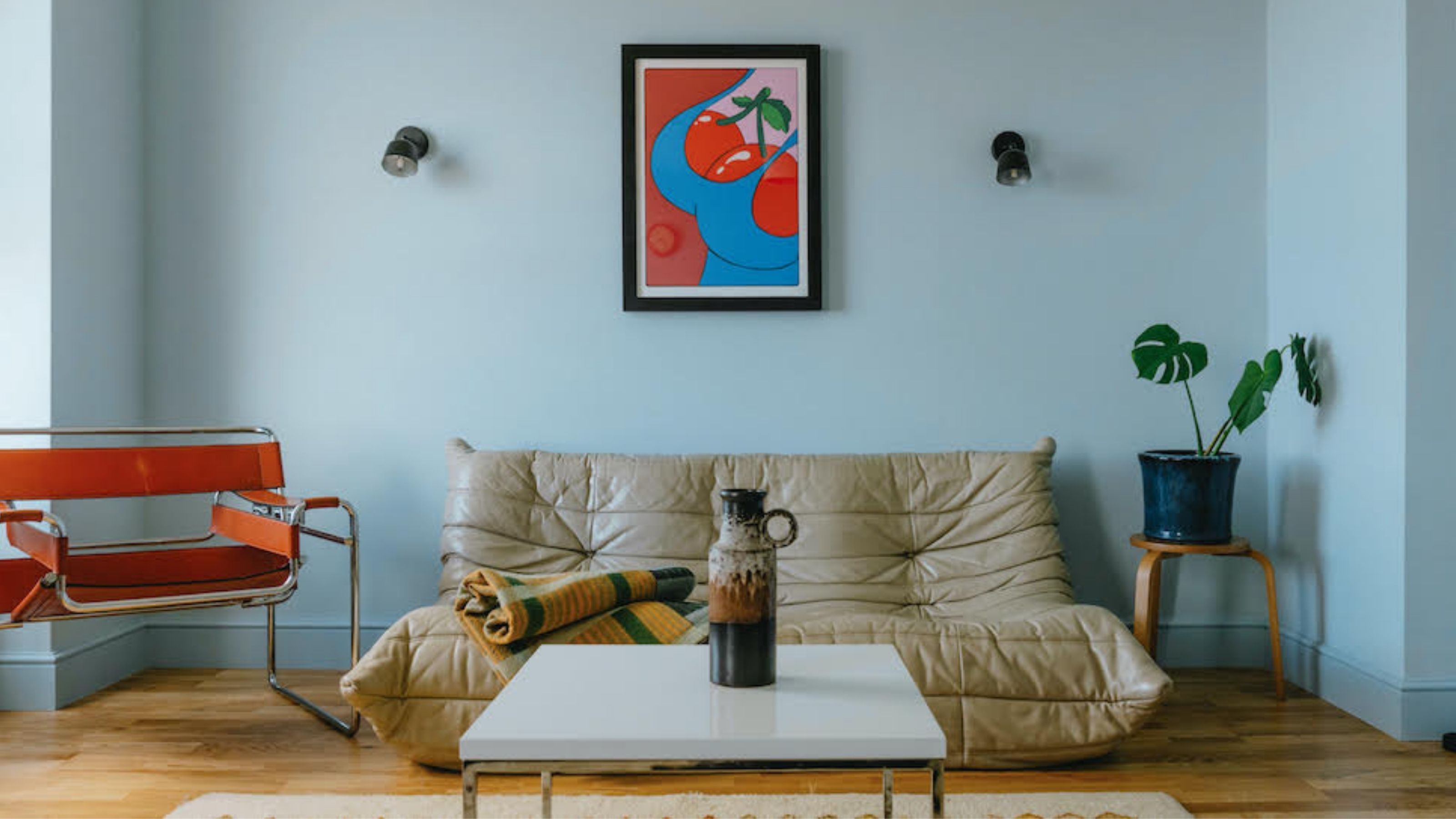 A light blue living room with a black-framed poster in the center of the wall above the beige leather sofa, with wooden flooring, a red metal-framed armchair and a plant in the corner on a stand