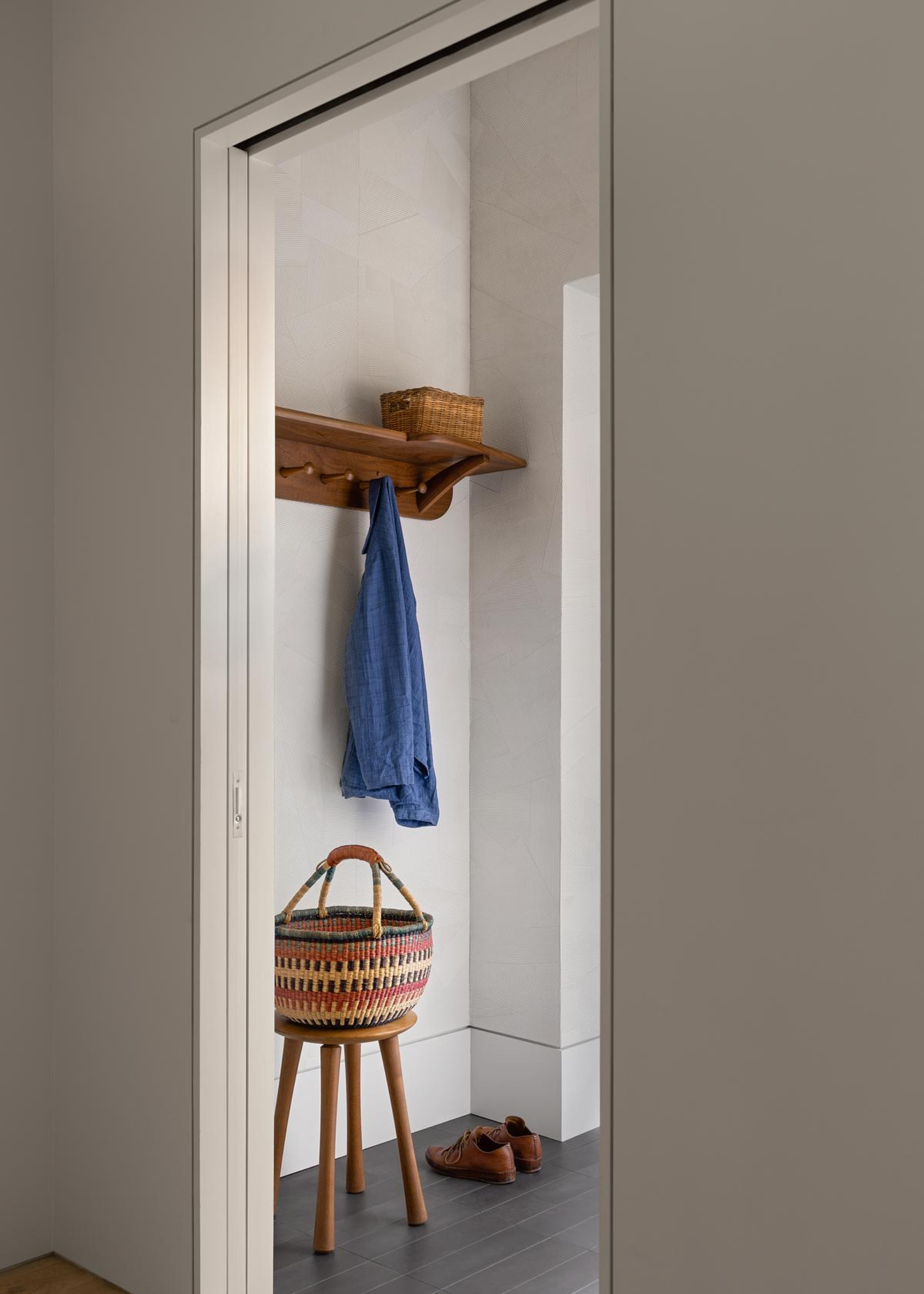 An entryway with coat hooks and a coat hanging by it. There is also a stool with a basket on top of it, and shoes placed next to it.