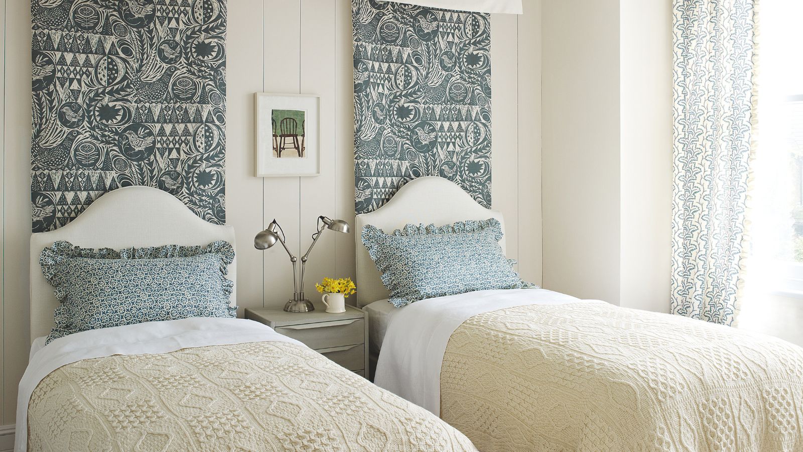 A white panelled twin bedroom with two twin beds with white headboards, with matching cream blankets, scalloped patterned blue pillows, and patterned wall tapestries behind each bed. Above the panelling is a strip of dark green wall, with white cornicing above.