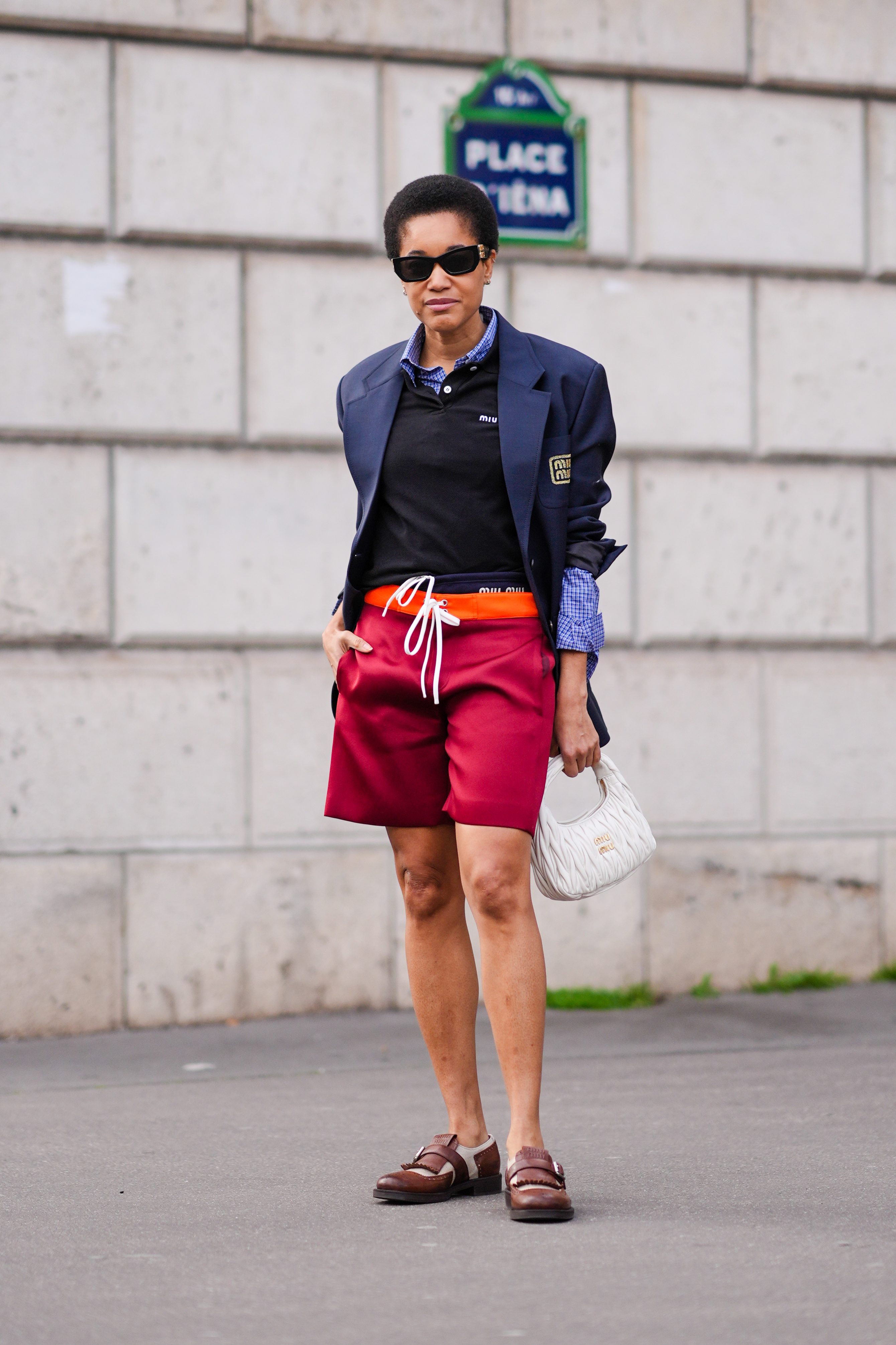 Tamu Mcpherson wears sunglasses, a blue shirt, a navy blue oversized blazer, a black neck top, red board shorts, a white bag, brown leather shoes at Paris Fashion Week on March 05, 2024.