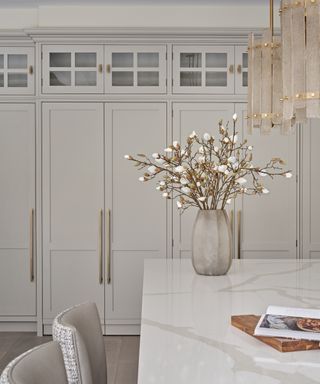 kitchen with off white/pale gray cabinetry with glazed doors at top, marble countertops, modern pendant lights, bar stools, vase with white magnolia