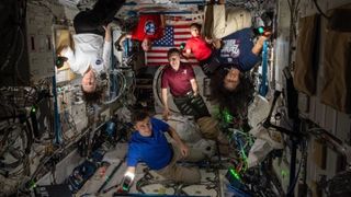 several astronauts in a dark module of the space station with their faces and bodies lit up by camera lighting. at back is an american flag