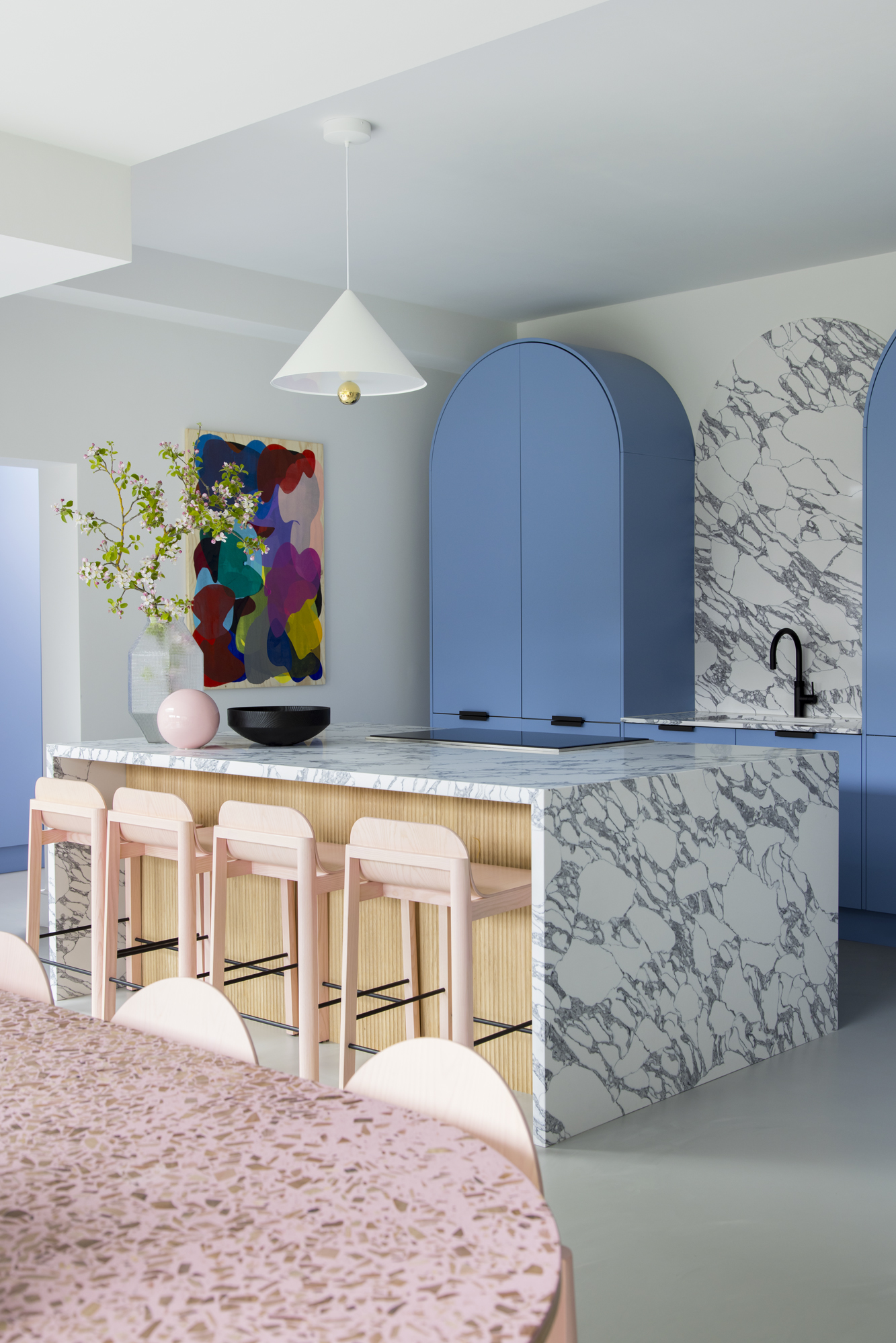 colorful kitchen with blue curved cabinets, marble backsplash and countertop, timber kitchen island with pink bar stool, a pendant light, bowls and vases on the bench with foliage and a large colorful artwork
