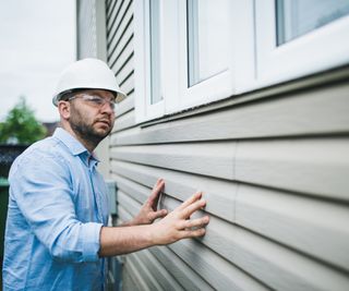 man with beard wearing white hard hat, safety goggles and blue shirt inspecting outside of weatherboard cladded property