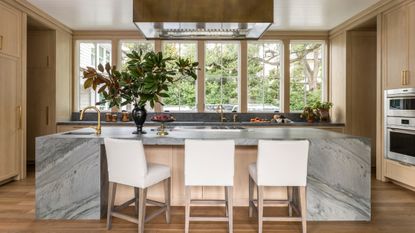 A wooden kitchen with a large stone island, where windows line the wall instead of a traditional backsplash
