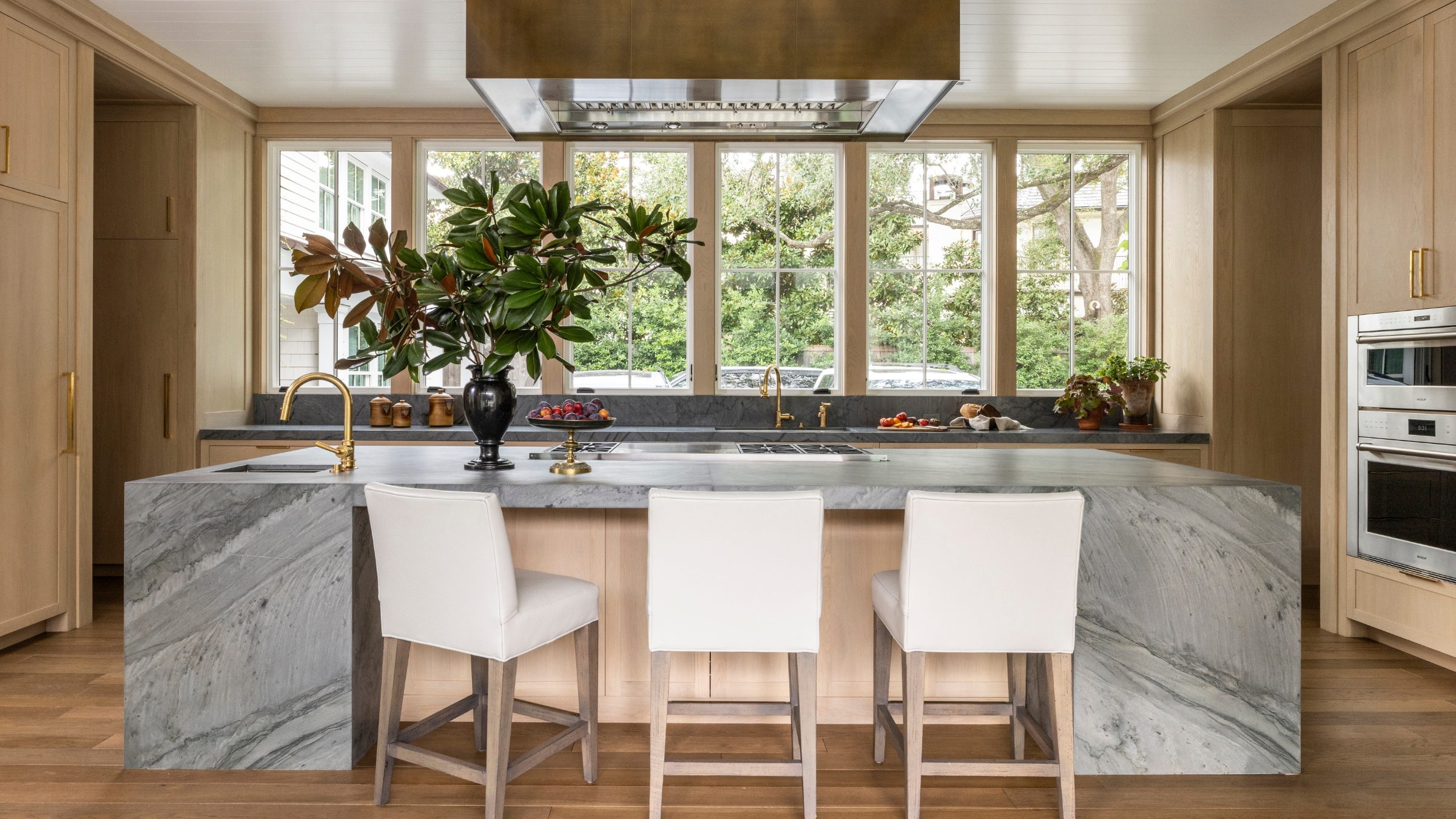 A wooden kitchen with a large stone island, where windows line the wall instead of a traditional backsplash 