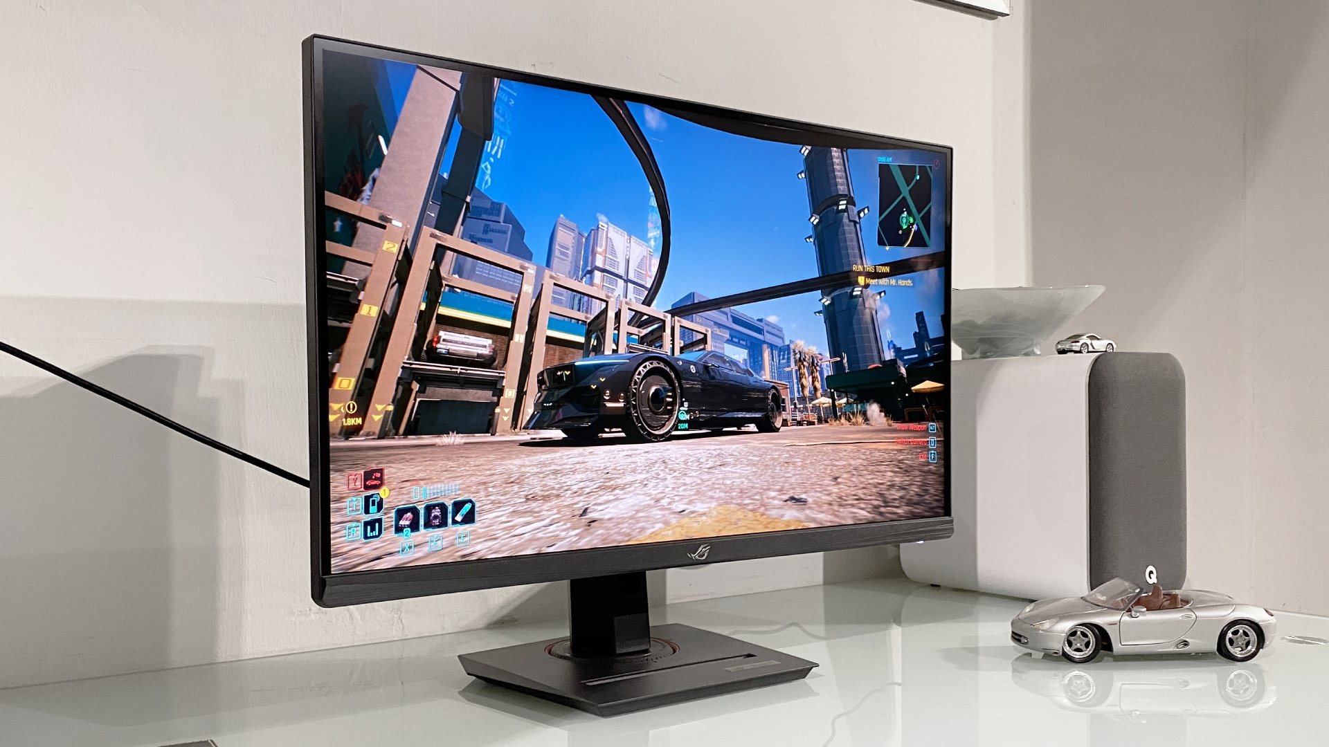 A sleek gaming monitor displays a vibrant cityscape from a video game. Next to it, a model car and speaker sit on a white desk, creating a modern tech vibe.
