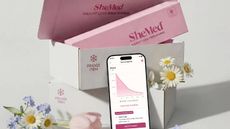 SheMed box containing SheMed products, with a phone displaying the SheMed app, surrounded by flowers. 