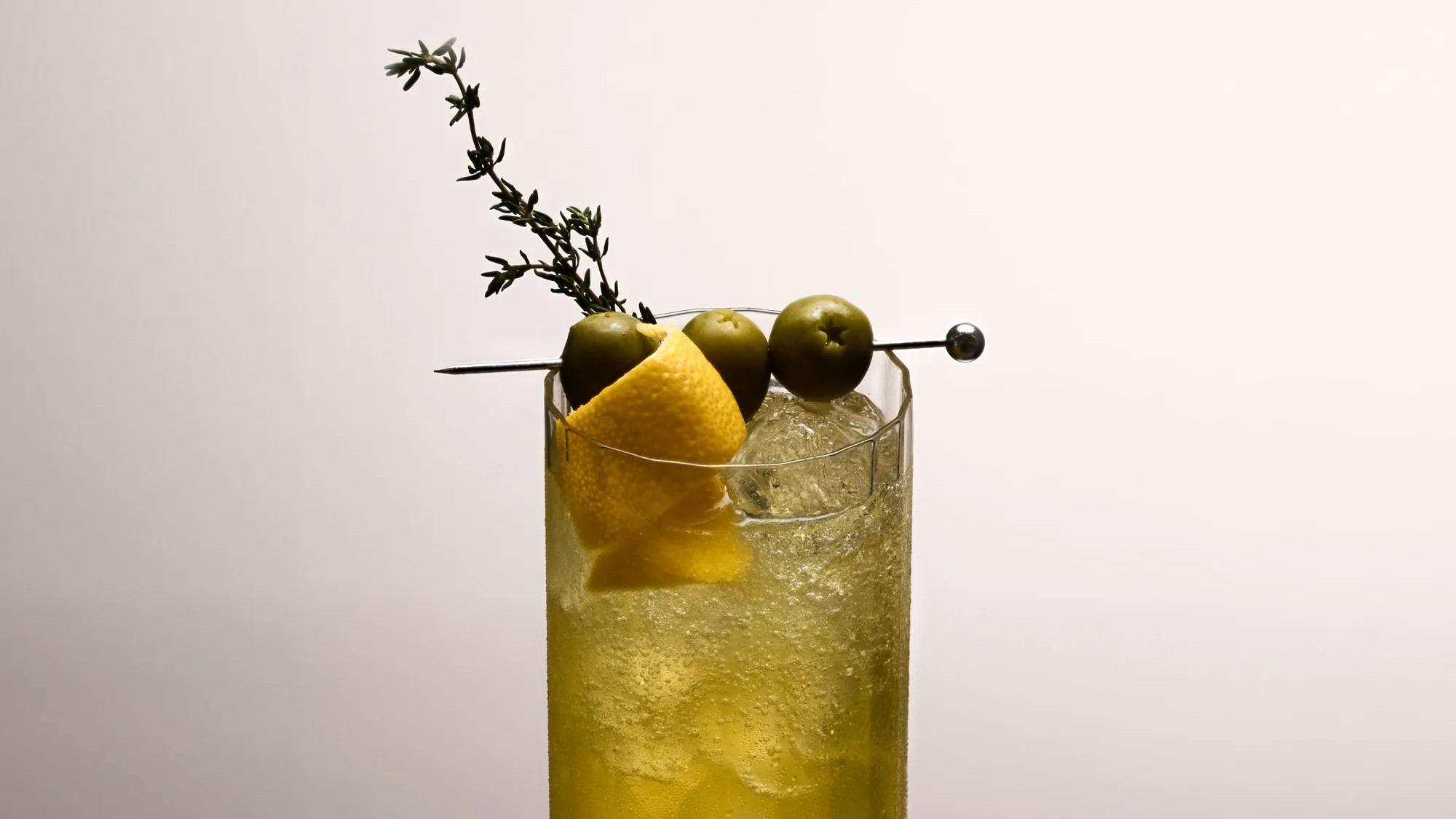 side shot of the top third of a collins glass filled with a dirty spritz and garnished with an olive skewer, thyme sprig and a lemon peel
