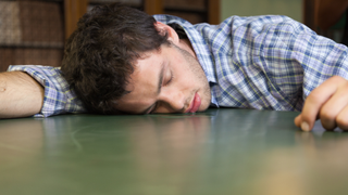 A man wearing a checkered shirt sleeping on a desk with a pool of drool next to his mouth