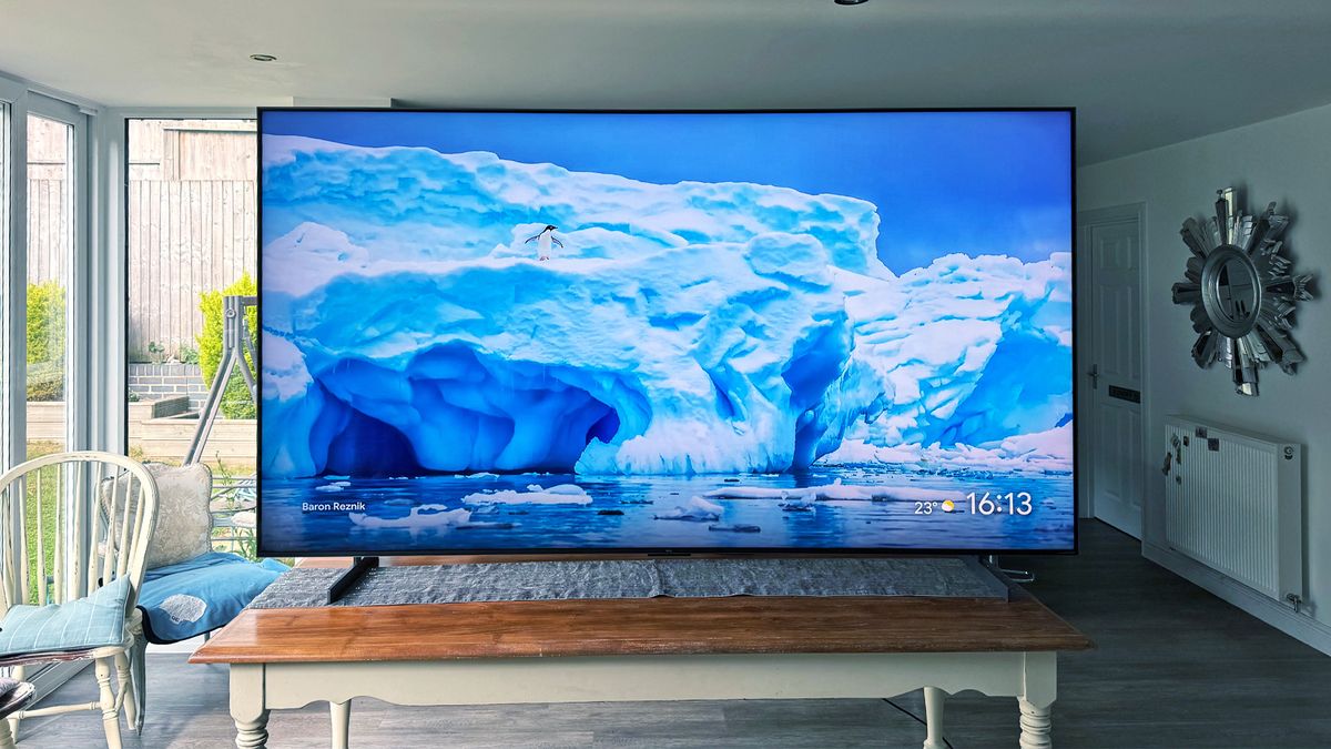 The TCL 98C7K 98-inch TV on a table in a living area, seen straight-on. On screen is a polar ice cap with a penguin just over halfway up it. This is our pick for best super-sized TV.