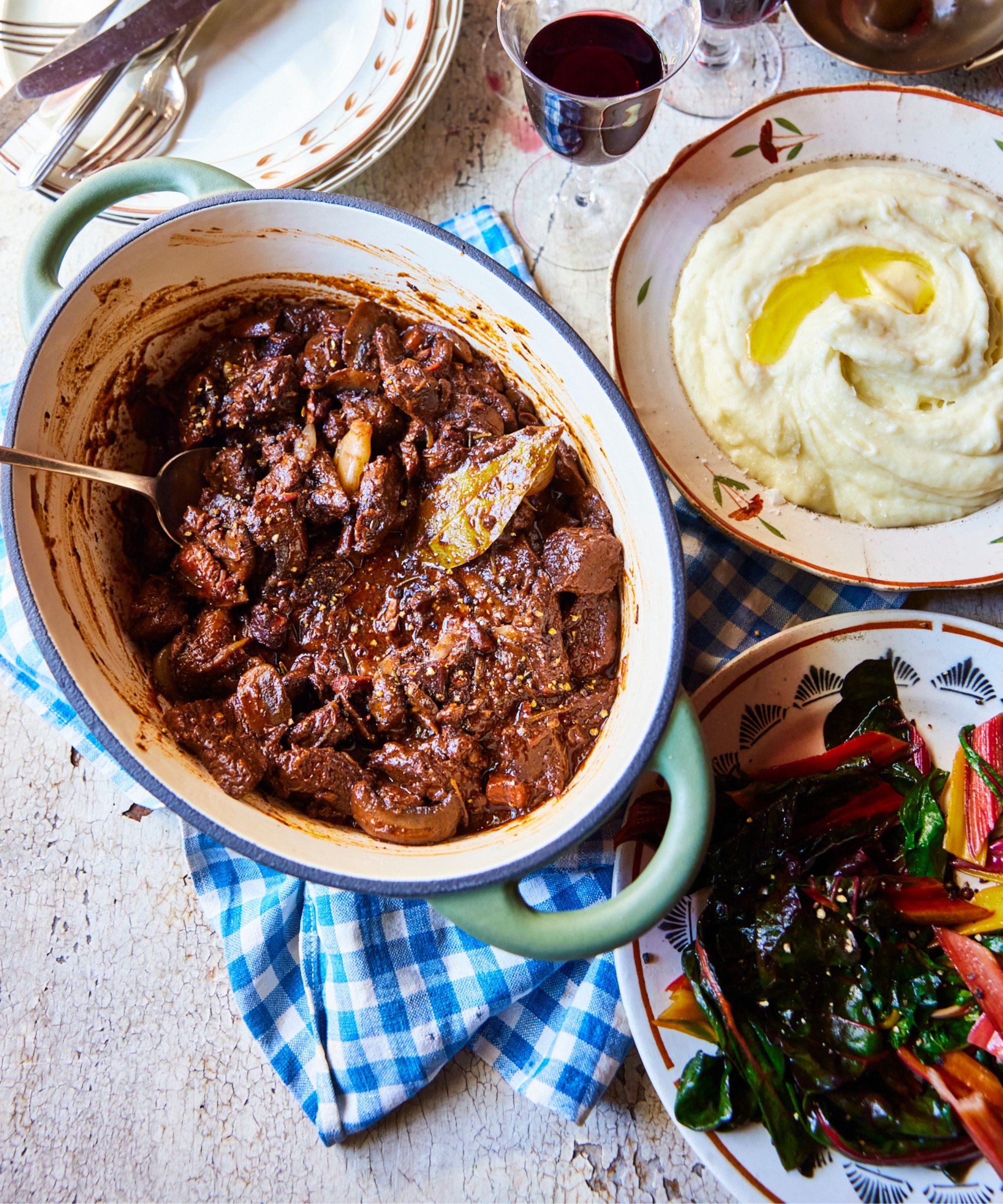 Pot of Beef bourguinon in a dutch oven on a blue gingham teatowel, next to bowl of colorful greens and another bowl of mashed potato