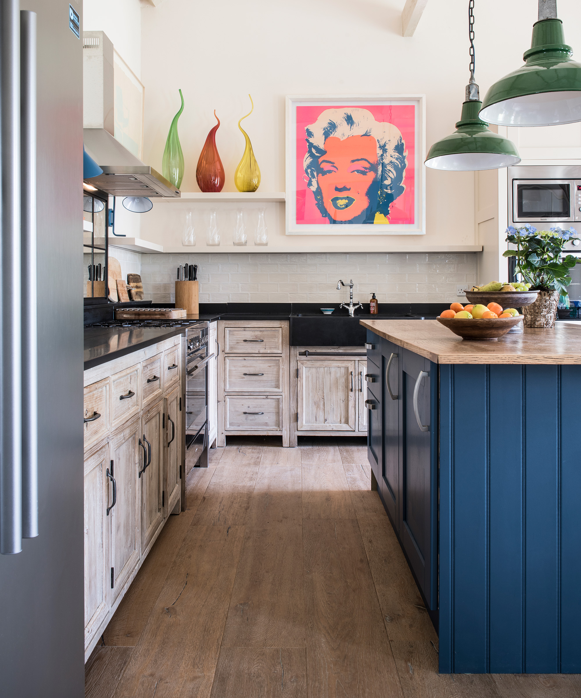 A kitchen with distressed wood cabinets and a blue, wood paneled island in front of green, red and yellow glass ornaments and a Marilyn Monroe artwork