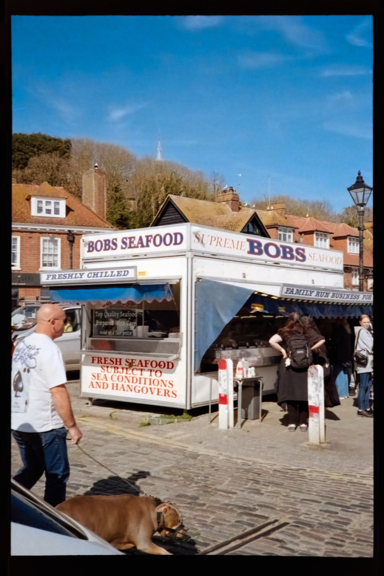 Scanned color negative of a seafood stall labeled &ldquo;Bob&rsquo;s Seafood,&rdquo; with customers gathered outside and a dog crossing the foreground.