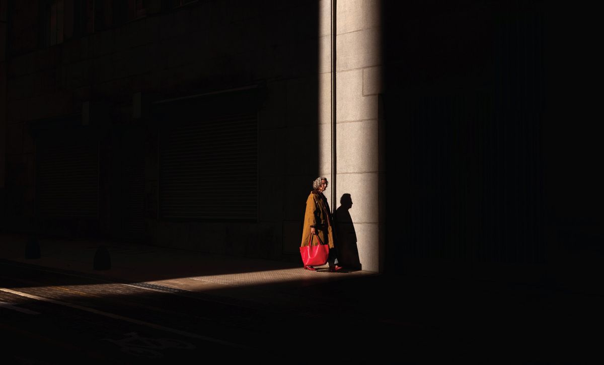 An elderly lady with a red bag standing in a beam of sunlight 