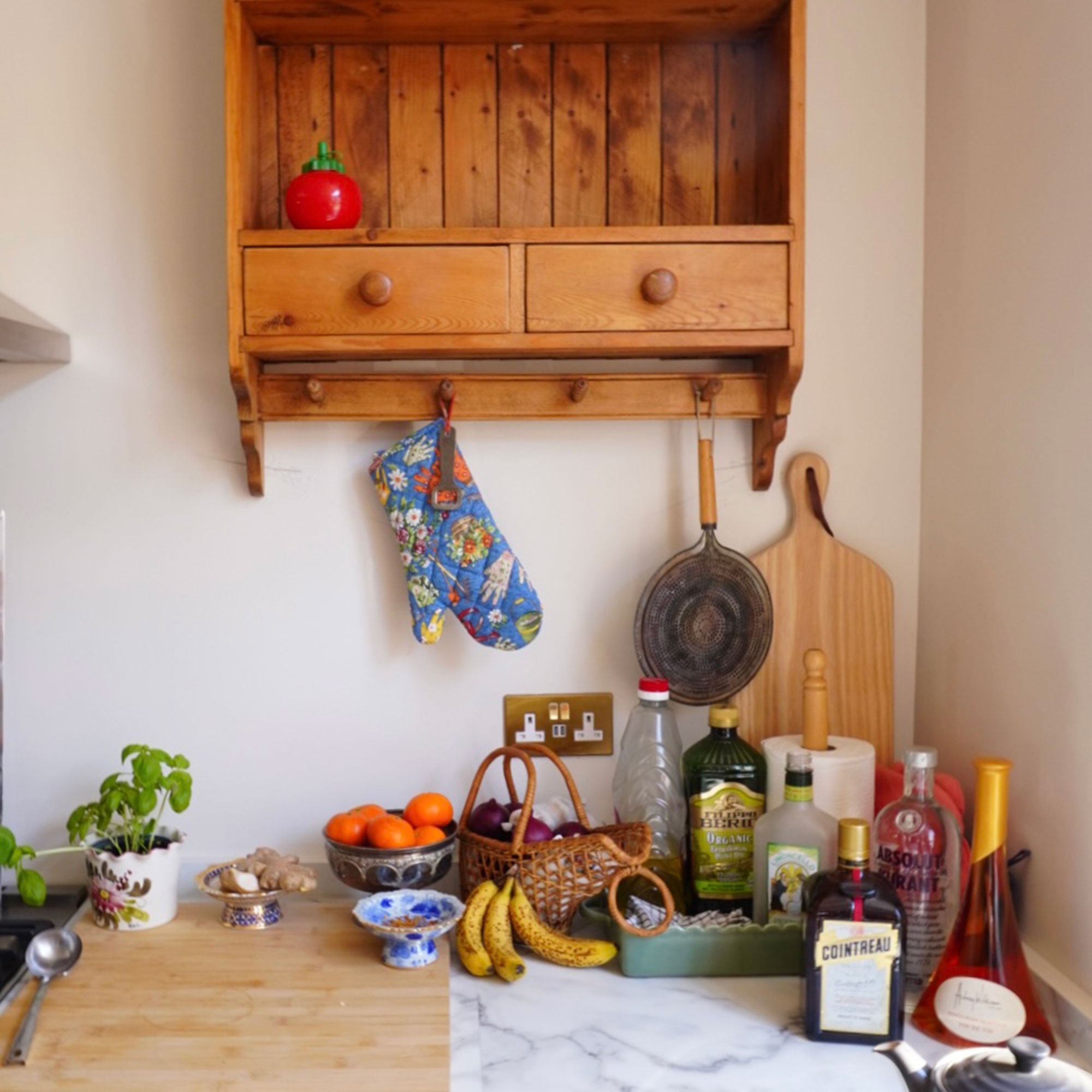 Corner of kitchen with wooden wall storage unit, and collection of bottles, chopping boards and fruit bowls on worktop