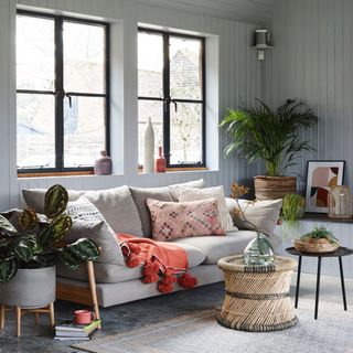 grey living room with tongue and groove walls, cream sofa, rattan side table, baskets, artwork, plants, textured cushions, rug, vases