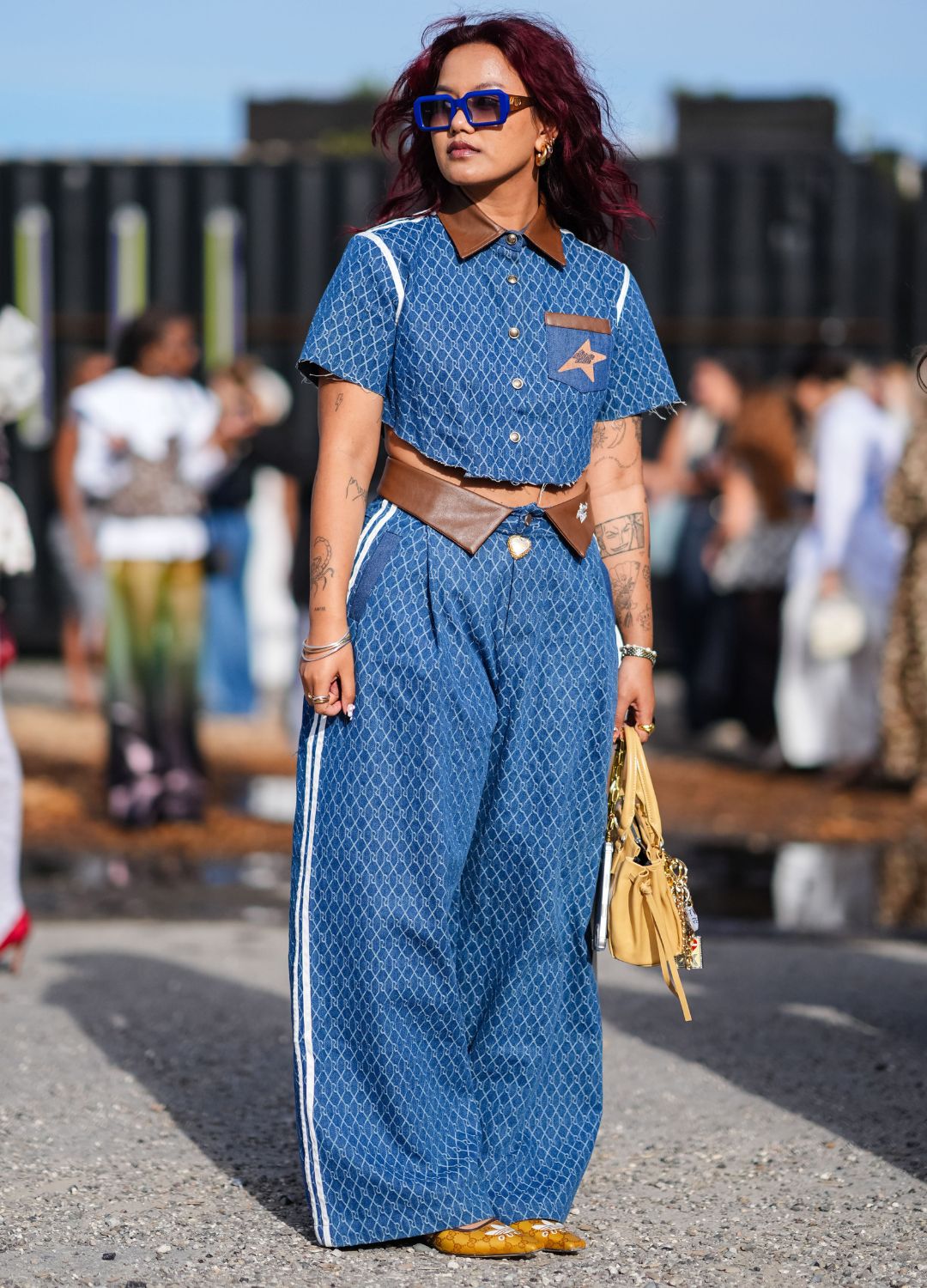A guest wears blue sunglasses, gold hoop earrings, light and dark blue Jacquard pattern short sleeve shirt with dark brown collar, light and dark blue Jacquard pattern baggy pants with white stripes and dark brown collared waist, silver bracelets, light yellow leather bag, Décolleté adidas x Gucci GG heels, outside Rolf Ekroth, during the Copenhagen Fashion Week Spring/Summer 2024-2025 on August 5, 2024 in Copenhagen, Denmark