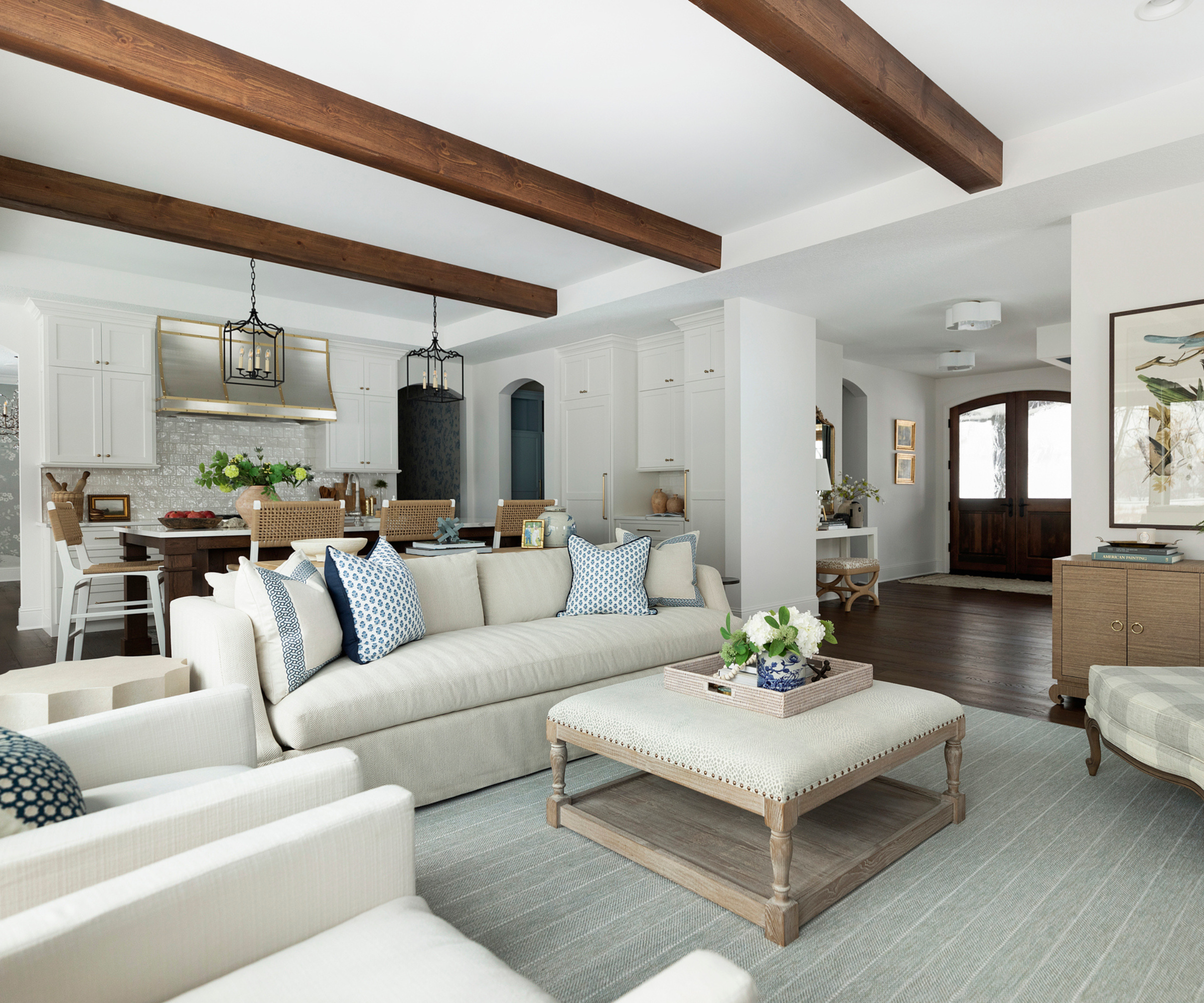 an open concept kitchen and living room with a neutral color scheme and blue accent decor, exposed beams on the ceiling, a couch, an ottoman, and a plaid accent chair in one corner