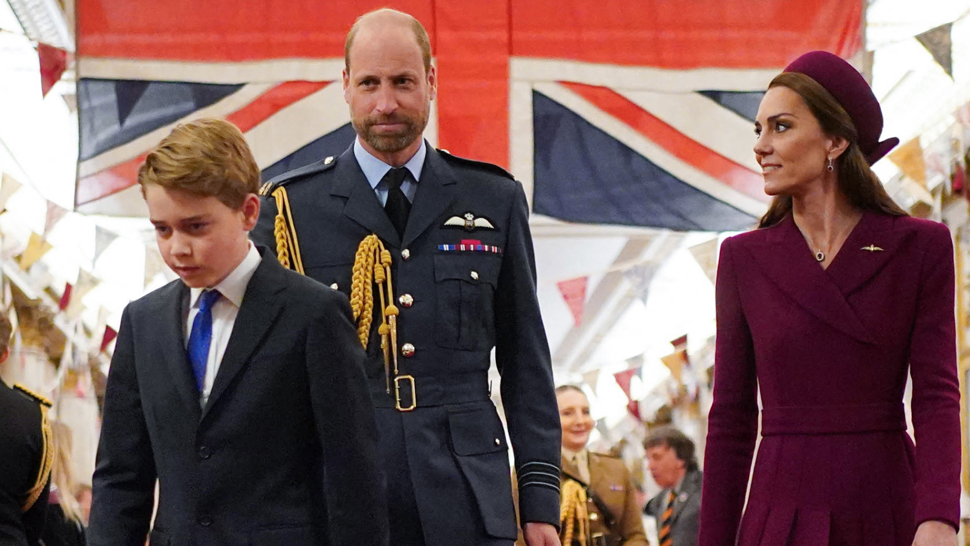Kate Middleton looks at Prince William, who is wearing military uniform in front of the British flag, while Prince George walks with his head down