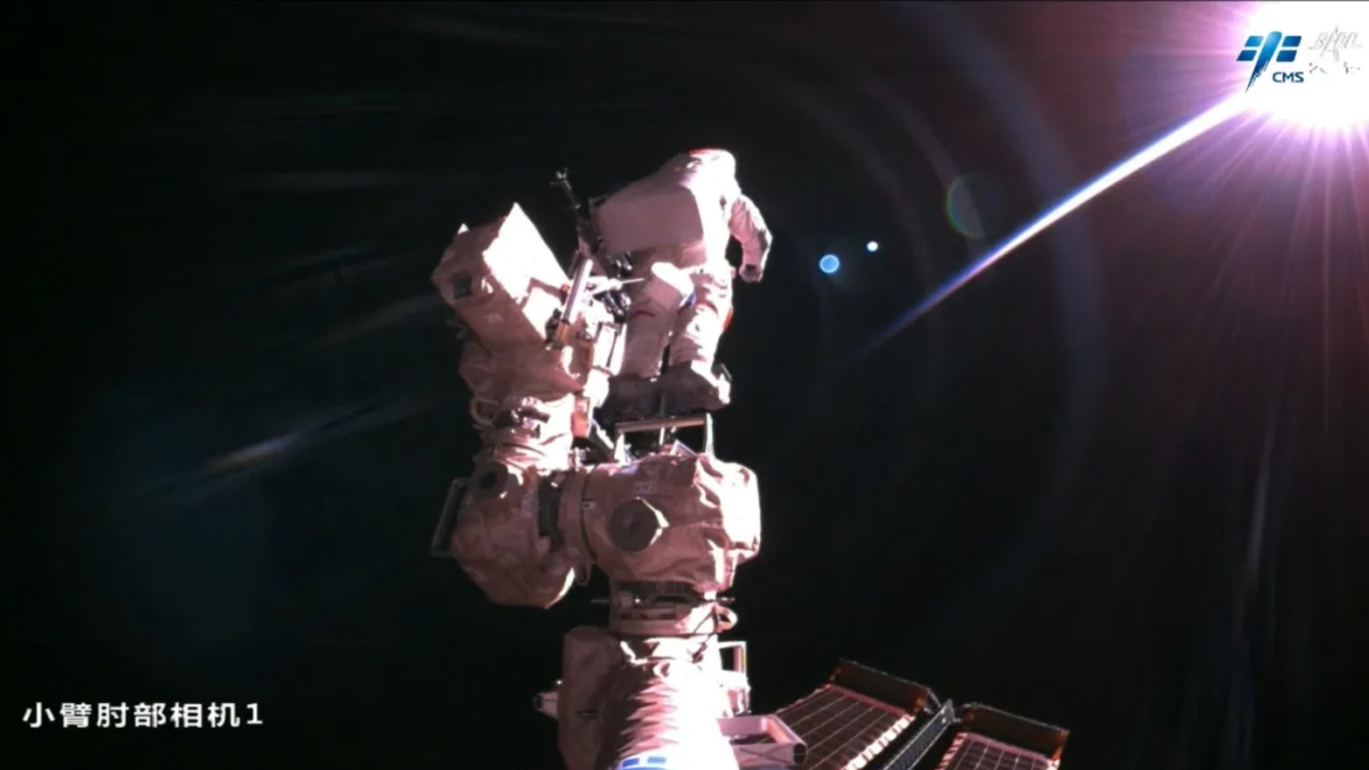 An astronaut wearing a white spacesuit stands at the end of a robotic arm with the blackness of space in the background