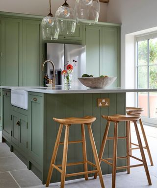 A traditional shaker-style kitchen with sage green cabinetry, a white Belfast sink, and a quartz worktop. Wooden stools and glass pendant lights add warmth and charm.