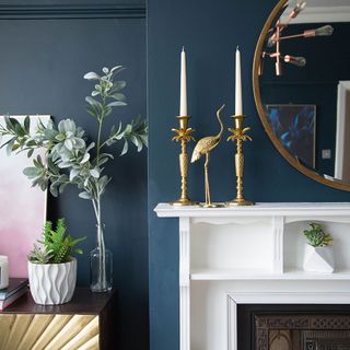 a close up of the white mantle around the fireplace in a living room with dark blue walls, with two gold palm tree candlesticks on the mantle alongside a gold bird statue, with pot plants sitting on a gold unit to the left